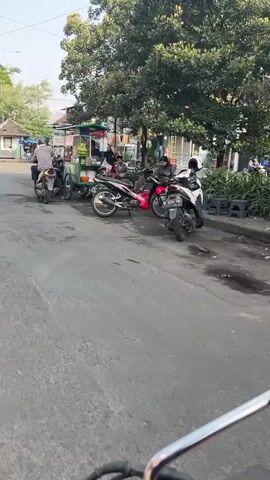 The video shows a street scene. Several motorcycles are parked on the side of the road. A food cart is set up, with at least two people attending to it. A few other people are seated near the food cart, some appearing to be customers. Trees line the street, and buildings are visible in the background. The overall atmosphere appears to be casual, possibly a local market or street food area.