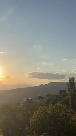 The video shows a sunset over a mountainous landscape. The sun is bright and slightly obscured by clouds, casting an orange glow across the sky. Below, a range of hills and mountains are visible in silhouette. A few buildings are nestled among the trees in the foreground, indicating a small village or settlement. The overall impression is of a serene and picturesque natural scene.