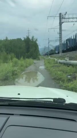 The video shows a view from inside a vehicle traveling on a dirt road with several large puddles. To the right of the road, there is a train track with a train passing by, and overhead power lines. Lush green vegetation lines both sides of the road and the train tracks. In the second frame, the vehicle drives through a large puddle, creating a significant splash of water.