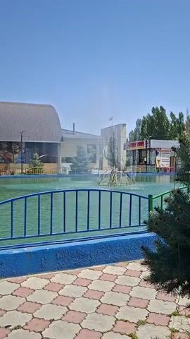 The video shows a view of a water park on a sunny day. In the foreground, there is a paved walkway with a pattern of pink and beige tiles. A blue barrier with a green fence runs along the edge of the walkway. Beyond the fence is a pool of turquoise water. In the background, there are buildings with pitched roofs and windows. Some of the buildings have large murals on their walls. A fountain sprays water in the middle of the pool. Trees and a blue sky are visible in the background.