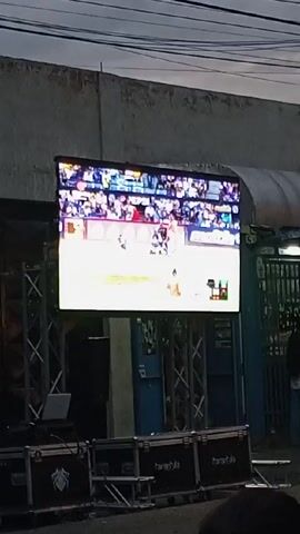 Here's a detailed description of the video:

The video shows a gathering of people watching a baseball game on a large outdoor screen. The screen is propped up by a metal frame structure, with some equipment boxes sitting beneath it. The baseball game appears to be in progress, with the pitcher in the middle of throwing the ball.

The camera zooms out, revealing a crowd of mostly men standing and sitting on chairs, motorcycles, and the sidewalk. They appear engaged in watching the game on the screen. The surroundings suggest an urban or semi-urban environment, with buildings visible in the background and overhead power lines extending across the frame. The sky is overcast.