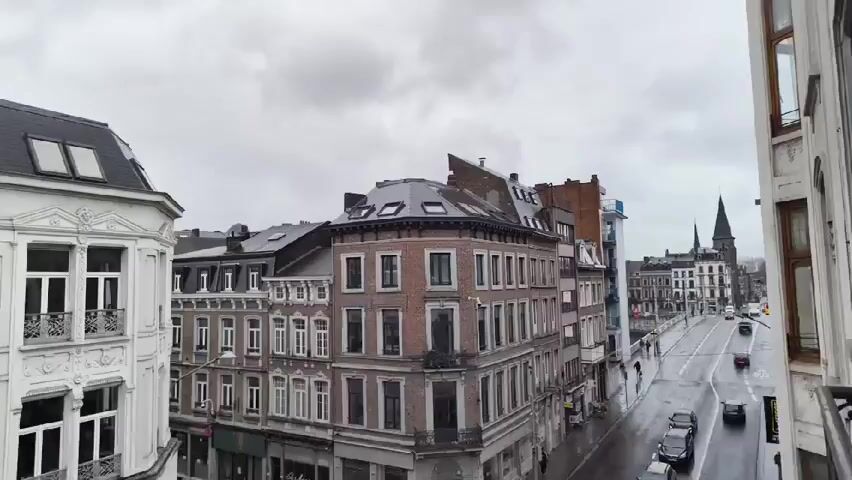 The video shows a street view in a European city, likely on a wet or overcast day. The camera, positioned from an elevated viewpoint, pans around, capturing the architectural details of the surrounding buildings. The buildings are multi-story with varying facade colors and styles, including white, red, and brown brick. Some have decorative elements like wrought iron balconies and ornate window frames. There are shops and businesses located on the ground floors of some buildings. People can be seen walking on the sidewalks and crossing the street. There are trams or trolleys running on tracks in the street. The overall impression is of a bustling urban environment with a mix of old and newer architecture.