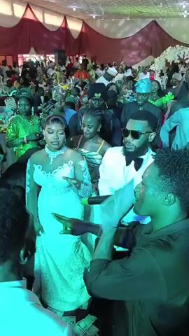 Here is a description of the video:
The scene is a vibrant wedding reception. A bride in a white lace dress stands beside a man in a white suit and sunglasses, both smiling as a man with dreadlocks, wearing a black shirt, sings into a microphone in front of them. The room is packed with guests, many dressed in traditional African attire, and most of them are holding up cell phones, presumably taking photos or videos. The venue is decorated with white and red fabric. There's a large floral arrangement in the background. The lighting in the room is bright and festive.