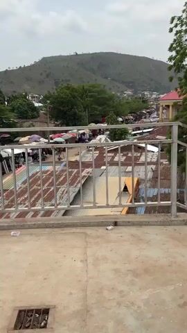 The camera pans around a busy marketplace scene in an unnamed town from a slightly elevated position.  The view is partially obstructed by a steel railing.  Below are corrugated metal roofs, many showing signs of wear and corrosion.  Further down, the marketplace is visible with numerous stalls covered by umbrellas and tarps.  People are milling about in the market.  The environment includes trees scattered throughout the town, a hill in the background, and buildings, some with red roofs.  A discarded pack of cigarettes lies on the concrete in the foreground.  The camera then pans to the right, moving down a set of stairs.  The marketplace is visible below the stairs.