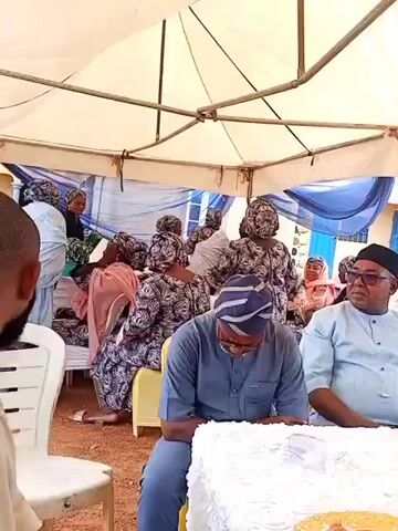 An outdoor gathering takes place under a large white tent. In the foreground, a bearded man in a light shirt looks towards the camera from the left. Next to him, a man in a blue agbada and striped cap sits, looking down at a white object on a table. To his right, another man in a light blue traditional outfit and dark glasses is seated, looking right. Behind them, numerous women are seated and standing, predominantly wearing patterned traditional Nigerian attire, including headwraps. Some women are seen standing and subtly moving, possibly dancing to the audible music. A building with blue walls is visible in the background, and the ground is reddish-brown earth.