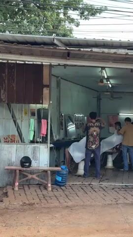 The video shows a barbershop with three barbers and at least two customers. One barber, wearing a dark shirt with a graphic design, is cutting the hair of a customer seated in a white barber chair, draped with a white cloth. Another barber, in a tan shirt and blue jeans, is standing behind a second customer, also in a barber chair with a white cloth. A third barber, wearing a white shirt and jeans and a mask, appears to be assisting or waiting. The barbershop is open-air with a corrugated metal roof supported by wooden beams. Shelving with towels and hair products is visible on the left. A water jug sits on the floor near the entrance. A small wooden bench with a black helmet on it is in front of the shop. The ground in front of the shop is dirt, and a paved area with bricks is present. Trees and power lines are visible in the background.