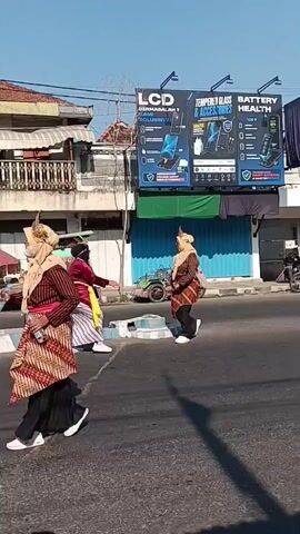 A group of people in traditional clothing are walking in a street. The people are wearing long skirts, blouses, and headscarves. They are also wearing patterned sashes around their waists. The people are walking in a line, and some of them are carrying water bottles. The street is paved with asphalt, and there are buildings on either side of the street. There are also some trees and plants in the background. The sky is blue, and the sun is shining. The video appears to be filmed on a bright, sunny day.