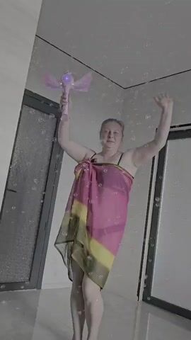 A person stands in a room that appears to be a bathroom. They are wearing a pink and yellow sarong over what looks like a black swimsuit. The person is holding a pink wand with wings and a glowing purple light on top in their right hand, raised above their head. Their left arm is also raised. There are white bubbles or sparkles in the air around them. The person's mouth is open as if they are speaking or singing. They are barefoot. To the left and right of the person are doors with frosted glass panels. The floor is light-colored and reflective. The walls are white. The lighting is bright.
