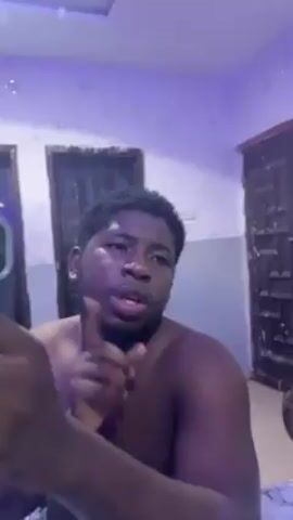 A shirtless man is indoors, recording a video. The room has two dark-colored doors, one of which is slightly ajar. The walls are painted white and the lighting changes from pinkish-purple to green. The man speaks, gestures with his hands, and touches his forehead. A smartphone is visible in his left hand.