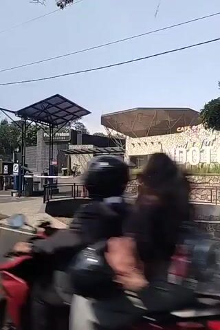 The video shows a road in front of a facility with an entrance gate. A person wearing a reflective vest is walking near the gate. There are signs indicating "Plant Protein" and "Indo Tofu". A billboard for "Indo Tofu Cafe & Resto" is visible. Several motorcycles are passing by, some with riders. The environment appears to be outdoors on a sunny day.