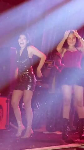 Two young women are dancing on a stage. The woman on the left wears a sparkly black mini-dress and clear heels. The woman on the right wears a red tank top and a black skirt with combat boots. The stage is lit by spotlights, creating a hazy, atmospheric effect. In the background, there are musical instruments and equipment, suggesting a performance. There is also a person in the background holding a camera or phone, possibly recording the event. The overall scene appears to be a lively event, possibly a concert or party.