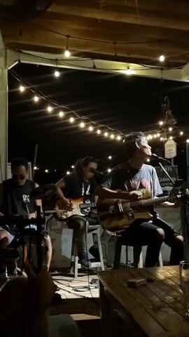 Here's a detailed description of the video:

The video shows a live band performing outdoors at night. There are three visible members, each seated on stools. The man in the center is playing an acoustic guitar and singing into a microphone. To his left is a man playing an electric guitar. The person to the far left seems to be playing a keyboard.

String lights are strung overhead, providing illumination, and a ceiling fan is visible in the top left corner.  A small wooden table is in the foreground, with a pint glass resting on it. In the background, there's a sign that mentions Guinness. The setting appears to be a bar or restaurant patio.