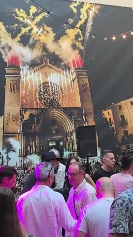 The video shows a crowd of people gathered at an outdoor event, likely a party or celebration, set against a backdrop depicting a church illuminated by fireworks. Most of the attendees are men dressed in casual summer attire, including button-down shirts, t-shirts, and one man is wearing a cowboy hat. Some are talking amongst themselves, while others appear to be looking towards the front where the large backdrop is placed. The backdrop is a large print of a church with fireworks exploding behind it, possibly signifying a special occasion or event. The event is being held in what appears to be a courtyard, as a stone wall is visible to the right, and string lights hang overhead, adding to the festive atmosphere. A speaker is set up in front of the backdrop, which suggests that music or announcements are part of the event.