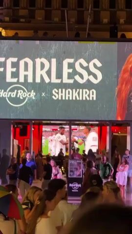 The video pans across a crowd of people gathered in front of a large outdoor screen at night. The screen displays the words "FEARLESS HARD ROCK X SHAKIRA" in large font, with a partial image of a woman with long, red hair on the right side. Below the screen is the entrance to a building with a red interior, where a live event appears to be taking place, with people visible on a stage or platform. The crowd is diverse, with individuals of various ages and ethnicities. Some people are looking up at the screen, while others are talking amongst themselves. The environment appears to be a public space, possibly a street or plaza, with buildings and seating visible in the background. The overall atmosphere seems lively and festive, with people enjoying themselves.