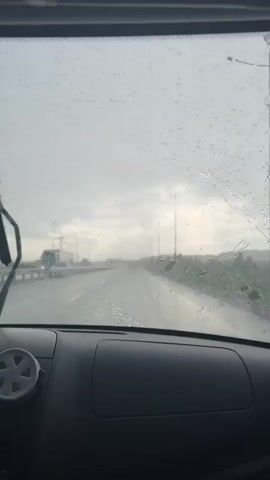 The video shows the perspective of someone driving in heavy rain.  The view is through the car's windshield, which is covered in raindrops, blurring the visibility. The windshield wipers are moving back and forth, attempting to clear the water. The road ahead is wet and glistening, with streetlights lining the side.  Other vehicles are visible in the distance, including what appears to be a large truck to the left. The sky is overcast and gray, suggesting poor weather conditions. The car's dashboard is in the foreground.