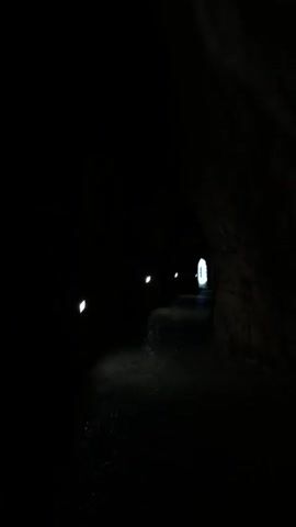 The video shows a dimly lit tunnel with rough, uneven walls. Small lights illuminate the path at intervals. The end of the tunnel is visible as a bright opening. The camera moves forward through the tunnel. The word "Minecraft" is spoken.