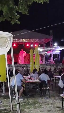 This video shows an outdoor evening event with a stage in the background. A performer in a large, flowing pink costume is on stage, illuminated by red lights. A banner with text is visible above the stage. In the foreground, several people are seated at tables, seemingly watching the performance. Yellow umbrellas are positioned between the audience and the stage. The environment appears to be a festival or gathering, with stone walls and trees visible.