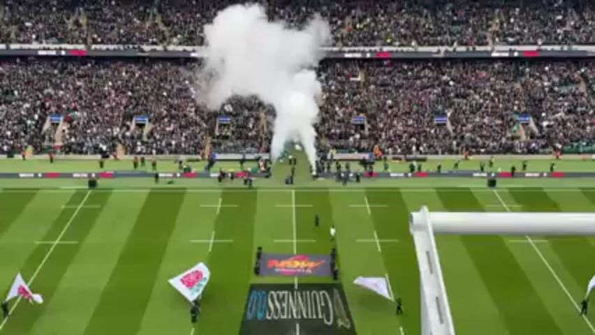 The video shows a rugby stadium packed with spectators.  Smoke cannons are firing as a team in green uniforms runs onto the field, likely for the start of a game. Flags bearing a rose emblem are visible on the field, along with banners for sponsors, including Guinness.  The view is from a high vantage point, offering a broad view of the field and stands.