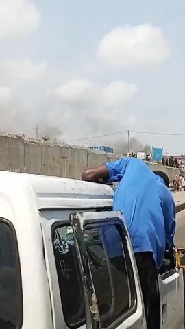 The video shows a street scene with several vehicles and people. In the background, there is a large concrete wall with some debris and what appears to be smoke rising from behind it, indicating a possible fire or disturbance. Several people are walking along the sidewalk and crossing the street. One person is sitting on top of a white van. Another white van and a yellow van are visible on the road. The sky is cloudy. There are various signs and stickers on the vehicles, including what appears to be military insignia on the white van in the foreground.