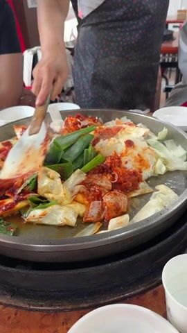 A person in a dark patterned apron is using a spatula to stir a dish of chicken and vegetables in a large pan. The dish appears to be Korean dakgalbi, a spicy stir-fried chicken dish. The pan contains pieces of chicken, cabbage, green onions, and a red sauce. The pan is on a stovetop or heating element. There are white bowls and cups visible on the table, and other people are seated around the table, though they are out of focus. The environment appears to be a restaurant.