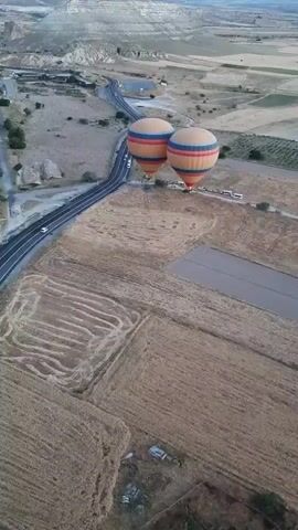 The video begins with an aerial view of two hot air balloons, one directly behind the other, floating over a rural landscape. Both balloons are colorful, with orange, yellow, and blue stripes. Below the balloons, a road with a few cars can be seen, as well as several agricultural fields. The landscape appears dry and arid, with rolling hills and distant mountains. The video then transitions to a wider aerial perspective, showing the hot air balloons continuing to drift over the landscape. The focus shifts to a single hot air balloon, which is closer to the viewer, and the camera pans to reveal more of the surrounding terrain. The video ends with a view of the landscape from the perspective of the hot air balloon, showcasing the patchwork of fields and a winding road below. The sky is overcast, suggesting it might be early morning or late afternoon.