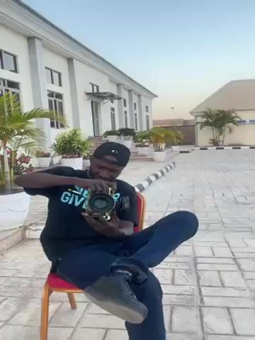 The video depicts a man sitting on a red chair in an outdoor setting. He is wearing a black t-shirt with the words "NEVER GIVE UP", blue jeans, and black sneakers. He also has on a black baseball cap. He is holding a camera up to his eye and appears to be taking a photo. Behind him is a large, white building with several windows and columns. There are also potted plants and palm trees in the background. The ground is paved with patterned concrete bricks. The sky is light blue, suggesting it is either early morning or late afternoon.