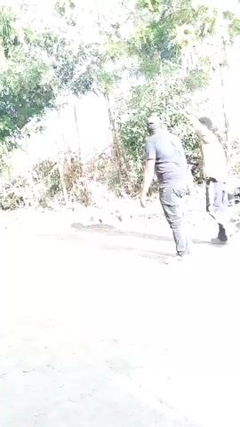 The video shows an outdoor scene on a sunny day with trees and foliage in the background. Two men are walking along a dirt road. One man is wearing a dark t-shirt, jeans, and a cap, while the other is wearing a tan jacket and dark pants with a stripe down the side. A woman in a white skirt, black t-shirt, and grey cardigan, carrying a shoulder bag, walks towards the camera. A motorcycle passes by on the road.