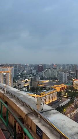 A high-angle view captures a cityscape at dusk. The camera pans from left to right, revealing a blend of traditional and modern architecture. Numerous buildings are illuminated with warm-toned lights, and the streets below are busy with moving vehicles.