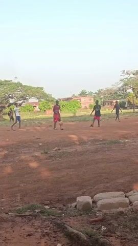 The video shows a group of young people playing soccer in a dirt field. There are five visible figures, all male, with several in athletic wear. They are spread across the field, kicking and chasing a soccer ball. The field is mostly red dirt with some patches of grass and scattered rocks. In the foreground, there is a pile of large stones. In the background, there are houses, trees, and a clear sky. The scene is captured from a static, ground-level perspective. The overall atmosphere is casual and active.