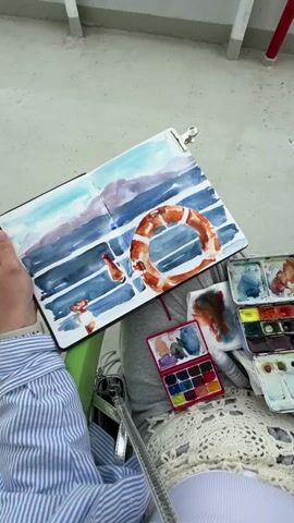 The video shows a person painting with watercolors on a ferry or a boat. The person is sitting down and holding a sketchbook on their lap. The sketchbook contains a painting of a life preserver attached to the railing of the boat, with the sea and land visible in the background. The person has several watercolor palettes and a brush on their lap. The boat has a white railing, a red fire hydrant, and an orange life preserver. The sky is cloudy, and the sea is calm. In the background, there is a town with parking lots and buildings visible.