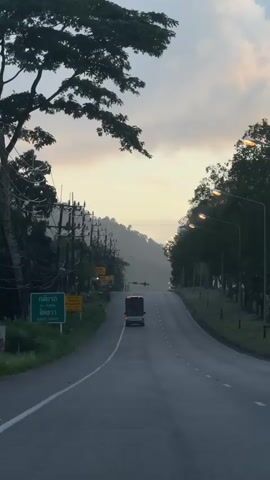 A van is driving down a two-lane road. The road is lined with trees and power lines. The sky is cloudy and appears to be during sunrise or sunset. There are some signs on the side of the road in a foreign language.