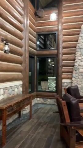 The video pans across a rustic, lodge-style interior. The walls are constructed from large, rounded logs stacked horizontally. A massive stone fireplace dominates one wall, adorned with a mounted bison head and a wooden mantelpiece featuring two lamps with orange shades and decorative log accents. Below the mantel, the fireplace itself is visible, appearing to have a modern electric or gas insert with faux logs. Two dark leather armchairs with wooden frames are positioned in front of the fireplace. To the left, a wooden table with drawers is set against the log wall, and large windows offer a view of an outdoor area with more stone and furniture. The floor is tiled with wood-look planks. The lighting is warm and ambient.