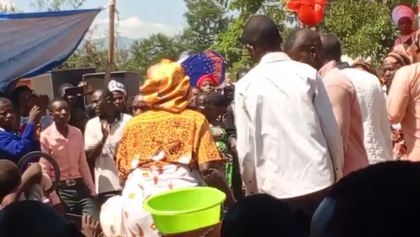 The video shows a large group of people gathered outdoors.  Most of the people are facing away from the camera, but it is possible to see that most of them are Black. They are dressed in a variety of clothing, including brightly colored prints, white shirts, and pink shirts. Some of the women have their heads covered with scarves. Red and blue cloth hang overhead as decorations. People are standing, and some are dancing.  The video seems to capture some kind of celebration.