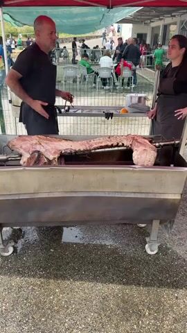 A whole pig is being roasted on a spit over an open flame. Two people, a man and a woman, are tending to the roast. The man, wearing a black t-shirt and apron, is holding tongs and appears to be basting or turning the pig. The woman, wearing a dark apron, stands beside him, observing. In the background, there are people gathered, some sitting at tables, under a tent. The setting appears to be an outdoor event or festival, with a fence and some trees visible. The ground is paved and appears slightly wet.