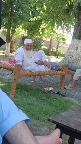 An elderly man with a long white beard and a white cap is sitting cross-legged on a traditional wooden charpai, resting his head on his hand. The charpai has a woven top with a colorful cushion and patterned pillows. The man is wearing a white shalwar kameez. He looks towards the camera, then adjusts his position. The scene is outdoors, with a well-maintained lawn and large trees in the background. A small wooden table is in the foreground to the right, and part of another person's blue shirt is visible in the lower left. Other people are partially visible around the edges of the shot. The ground around the charpai is covered with gravel. The setting appears to be a garden or backyard.