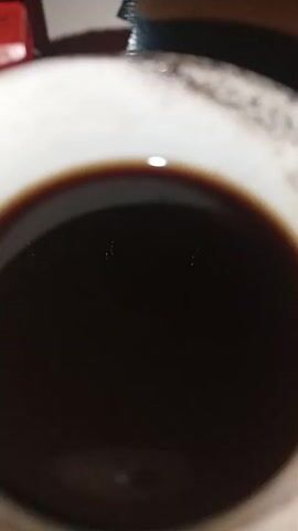A close-up, overhead shot shows a cup of dark liquid, likely coffee. The cup appears to be ceramic with a decorative rim. The liquid has a slight sheen, reflecting light from above. Two small, bright specks are visible on the surface of the liquid. The background is blurred but suggests an indoor setting.