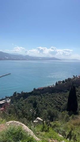 The video shows a wide shot of Alanya, Turkey, from an elevated perspective. A large cruise ship is docked at a pier extending into the turquoise sea. A medieval castle wall runs along the hillside on the right, amidst lush greenery and scattered trees. The landscape includes mountains in the background and a bright blue sky dotted with fluffy white clouds. As the camera pans slightly to the left, a brick structure is revealed. The foreground features rocky terrain and vegetation. The scene is bathed in sunlight, creating a vibrant and clear view of the coastal city and its surroundings.