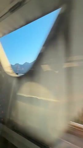 The video shows a view from a train window. The landscape outside includes a river, some vegetation, buildings, and mountains. The camera is moving, creating a blurred effect and reflections on the window. A person's hand is visible in the lower right corner of the frame, suggesting they are holding the phone recording the video. The sky is clear and blue. The time stamps indicate the video is approximately 4 seconds long.