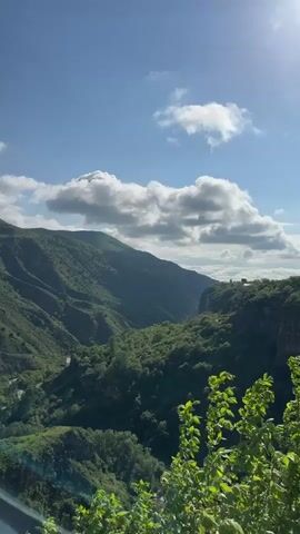 The video shows a scenic mountain range under a partly cloudy sky. The mountains are covered with green vegetation, and a river runs through the valley between them. The camera is relatively still and pans slightly across the landscape. A small building is visible on top of the cliffs, appearing to be at the far end of the canyon. The wind sound is prominent.