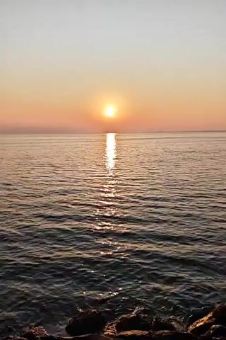 The video shows a sunrise over the ocean. The sun is a bright orange orb low on the horizon, reflecting a path of light across the water. The sky graduates from a light blue at the top to a pale orange near the horizon. The water has small waves. Some dark objects can be seen in the lower left, likely rocks or part of a shoreline. The camera is steady.