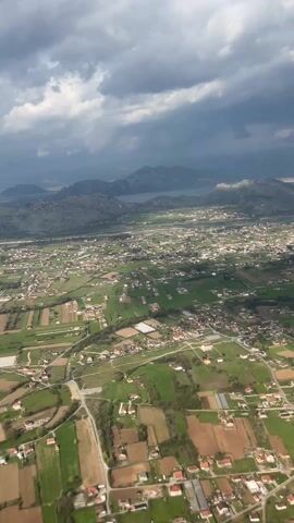 Here's a description of the video:

The video shows an aerial view from an airplane window of a rural landscape. Below, there's a patchwork of green fields and scattered houses, indicating a populated area. In the distance, there are mountains under a cloudy, gray sky, suggesting potential stormy weather. A portion of the airplane's wing is visible in the top right corner of the frame.