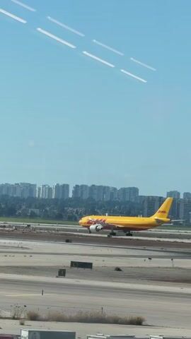A yellow and red DHL cargo plane is seen on an airport runway. The plane is in motion, with its landing gear down, appearing to be either landing or taking off. In the background, a city skyline with numerous buildings can be observed under a clear blue sky. The foreground shows the airport tarmac with various markings and some sparse vegetation. The lighting suggests it is daytime.