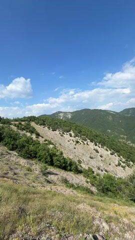 The video captures a panoramic view of a lush, green mountainous landscape under a bright blue sky with scattered white clouds. The foreground features rocky terrain with dry grass and some sparse vegetation. The camera pans slowly to the right, revealing rolling hills covered in dense trees and patches of lighter-colored rock formations. The perspective suggests the viewpoint is from a high elevation, offering a sweeping vista of valleys and distant mountain ranges. There are no people or discernible man-made objects visible in the scene. The overall impression is of a natural, serene, and expansive wilderness.
