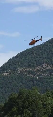 An orange helicopter flies through a blue sky with some clouds, with a long cable extending from it. A white container is attached to the end of the cable. The helicopter is positioned over a steep, tree-covered mountainside.