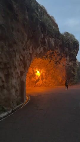 The video shows a person walking on a road near a tunnel. The tunnel is carved through a rocky cliff and is lit by a yellow light inside. The sky is overcast, indicating it's either early morning or late evening. The road is asphalt and has a curb on the left side. The person is walking away from the camera, towards the tunnel. The cliffs are textured and have some greenery on top.