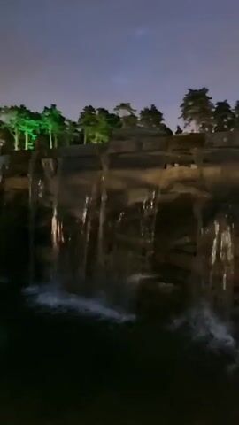 This is a low-light, static shot of a man-made waterfall at night. Trees are visible at the top of the waterfall, with some of them illuminated by green lights. The water cascades down over large rock formations into a dark pool below. The sky is dark with some faint clouds. The overall impression is one of a peaceful, illuminated natural setting.