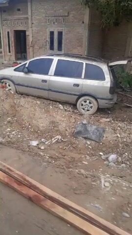 The video shows a dirt road, alongside a construction area with scrap metal, wood, and debris scattered around. Two abandoned or wrecked cars are visible. The first car is a silver station wagon with its trunk open. The second car is a dark red sedan, partially buried in the dirt. Beyond the construction area, there are palm trees and other vegetation. The overall scene appears to be a roadside view of a construction site in a tropical or semi-tropical location. There are no people visible in the frame.