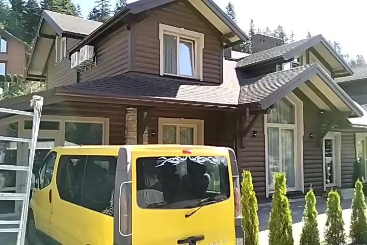 A yellow van is parked in front of a row of modern, dark brown houses with gabled roofs. The houses have numerous windows and are flanked by manicured evergreen trees. Beyond the houses, a paved road is visible, with several cars parked on the side. The environment is wooded, with tall evergreen trees forming a backdrop. The sky is blue with scattered clouds, indicating a daytime scene.