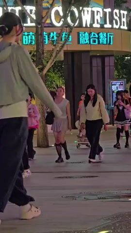 This media file captures a street scene at night, likely in the Tongzhou District of China, indicated by visible Chinese characters on a building facade. The primary focus is on a tall young woman in the foreground, wearing a grey hooded jacket over a white top and loose dark pants, with large beige platform shoes. Her hair is pulled back in a bun, and she wears glasses. She appears to be walking or dancing, with her body angled away from the camera.

Behind her, several other people are visible. To her left, a woman in a light-colored top and dark pants walks, and further left, a woman in a pink top and skirt is seen. To her right, a woman in a black and white outfit, resembling a maid costume, with thigh-high socks, walks with a pink backpack. A baby in a stroller is also present, pushed by a woman. Other individuals are in the background, suggesting a public area with pedestrian traffic.

A prominent building in the background displays a large sign with English text reading "THE COWFISH" and accompanying Chinese characters. The lighting suggests it is nighttime, with artificial lights illuminating the street and the building. The atmosphere appears lively, with various people engaged in activities. The ground is paved, with colorful illuminated lines suggesting a designated public space, perhaps for performances or gatherings. The overall scene depicts a bustling urban environment at night.