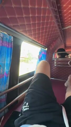 The video shows the interior of a bus or similar vehicle. The ceiling is padded with a diamond-quilted red material. Red padding with a similar diamond-quilted pattern lines the sides of the seats. Metal railings run along the aisle. A person's bare legs and feet are visible in the foreground, wearing black shorts. In the background, several people are seated, facing forward. The aisle is narrow, and there are multiple rows of seating on either side. Lights are visible on the ceiling. The overall impression is of a somewhat dated but well-maintained interior.
